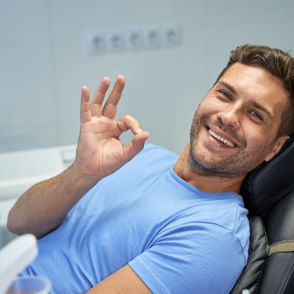 Contented young man showing an okay sign with his hand while sitting in dental chair and looking at camera